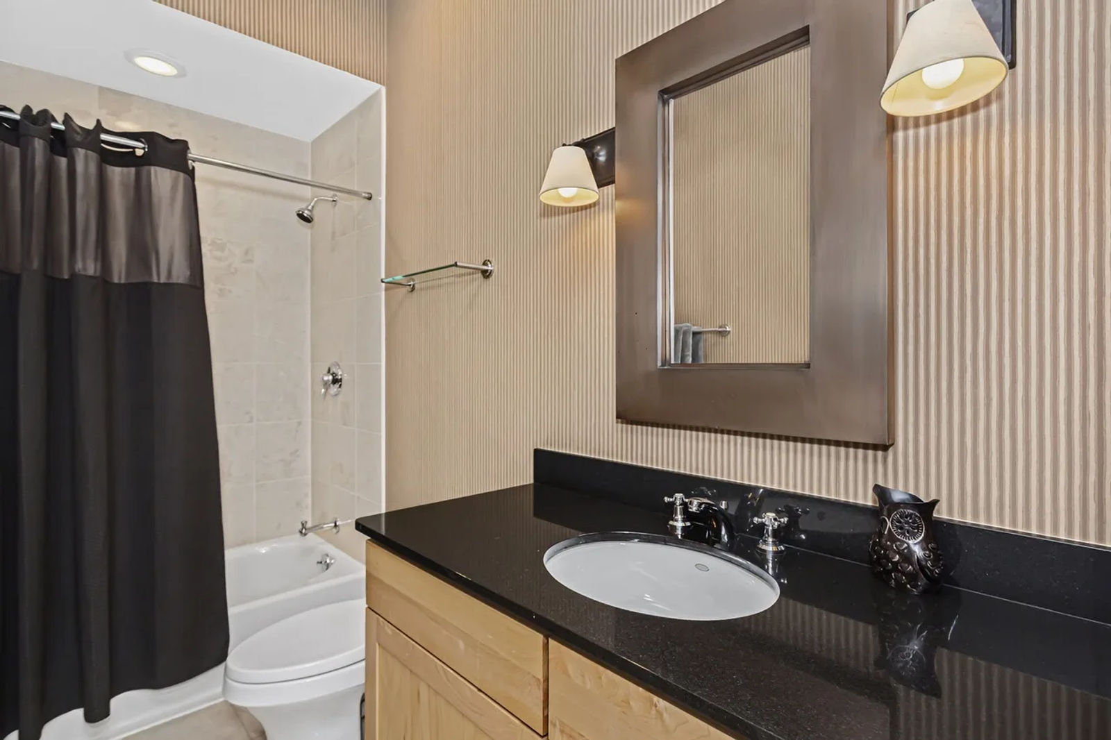 Before -- dated guest bathroom with tub and black curtain, maple vanity, black granite and striped wallpaper