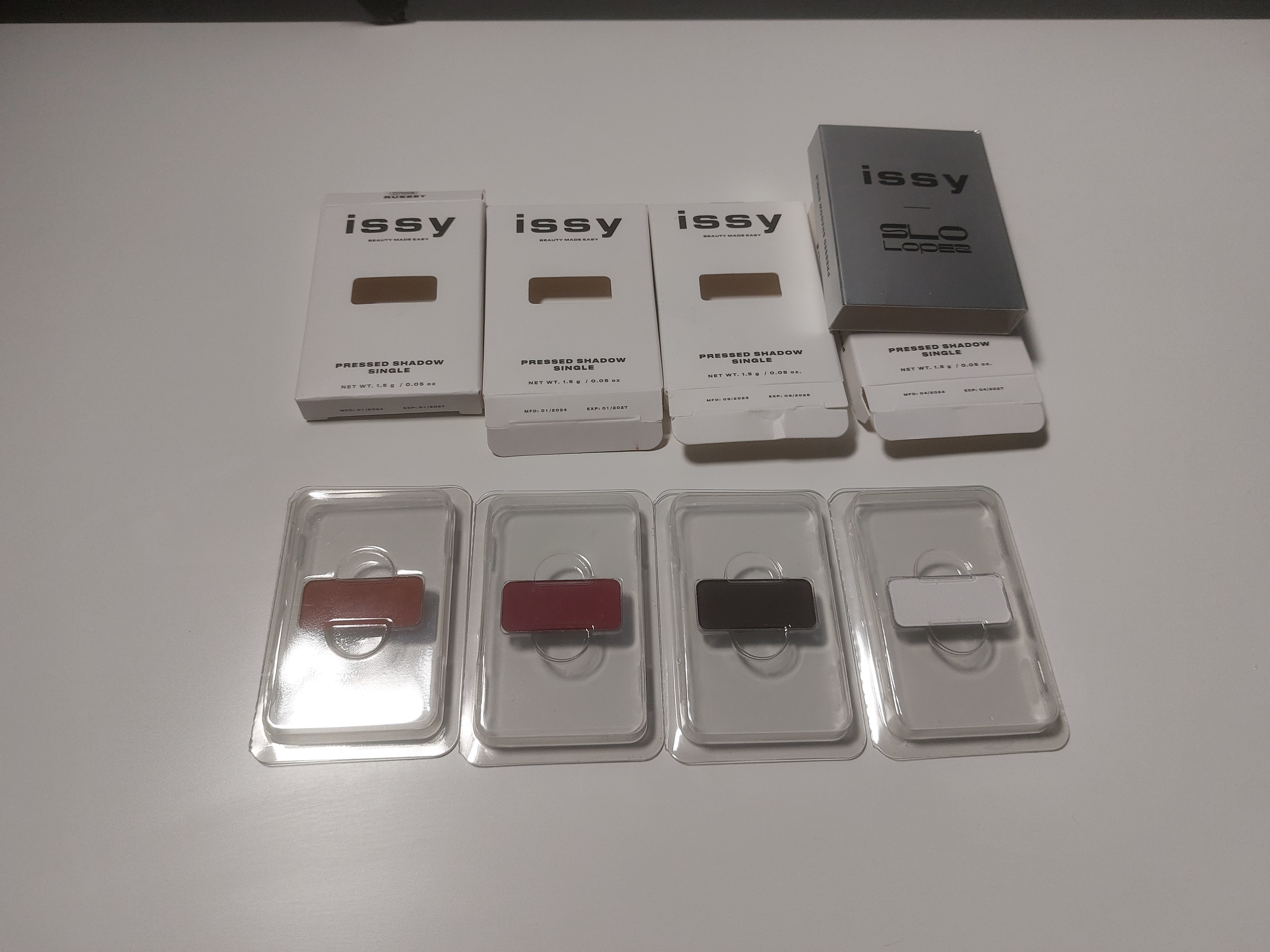 Four single eyeshadow pads in partially opened packaging. Each pad is displayed in a clear plastic container, and the colors appear to be red, dark red, black, and white. Above the pads, there are four white boxes labeled "issy" with the words "Pressed Shadow Single." One box is silver with "SLO Lopez" branding, also labeled as a pressed shadow. The products are arranged on a plain white surface.