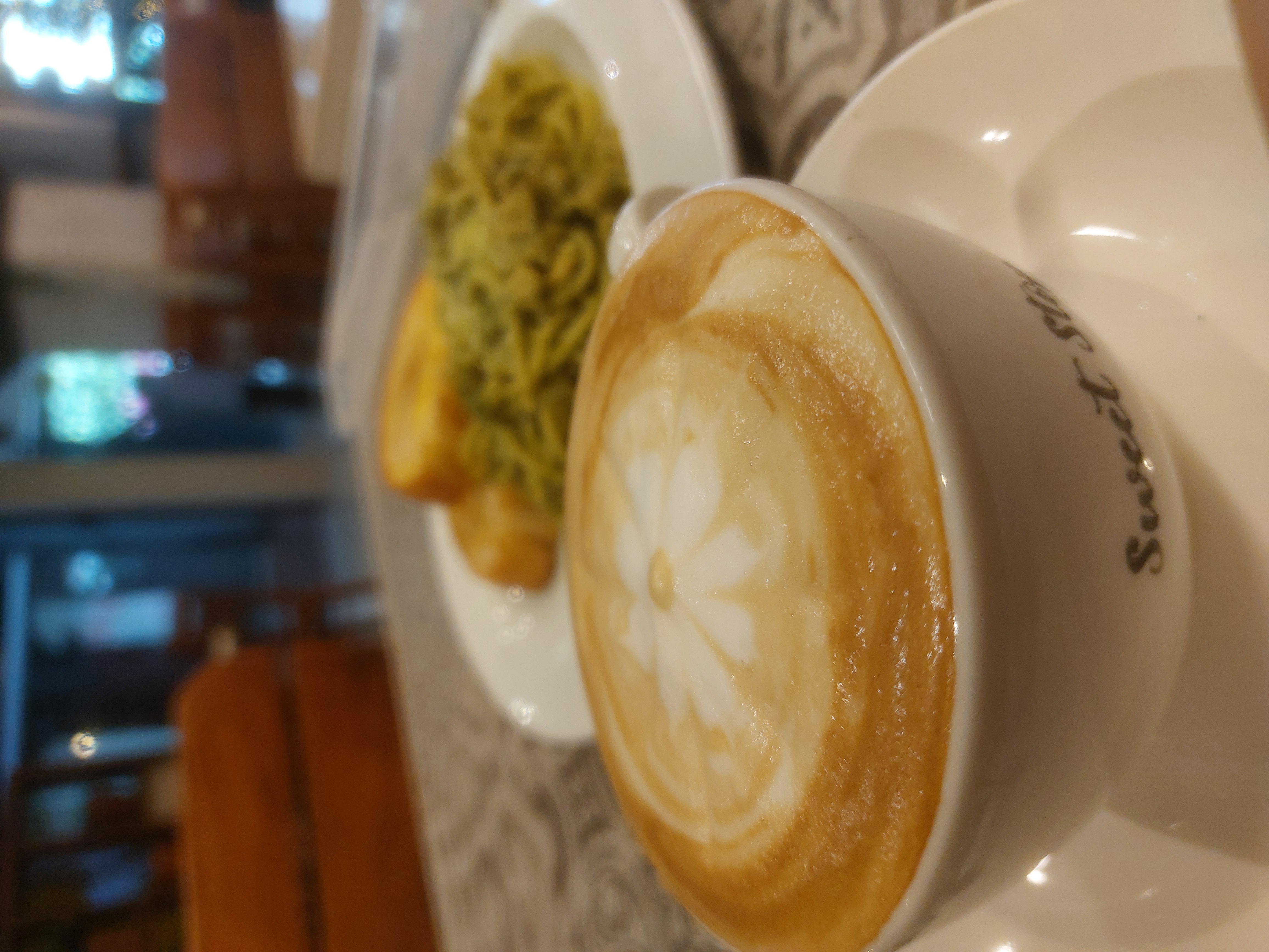 Coffee topped with milk foam and a floral design. In the background, a pair of toasted breads and pesto pasta on a white plate.