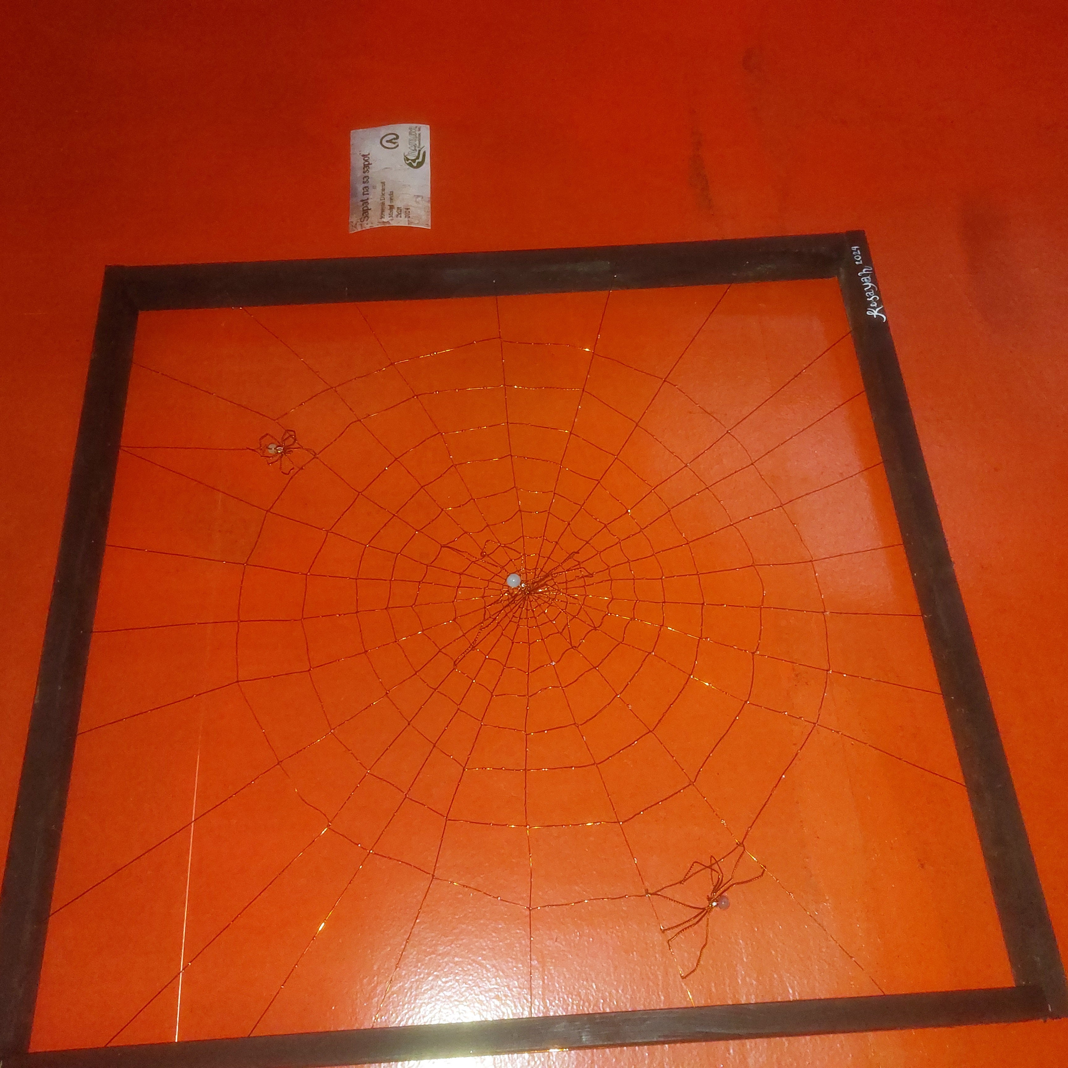 An artwork depicting three spiders on a web. It's made of copper wires inside a square wooden frame.