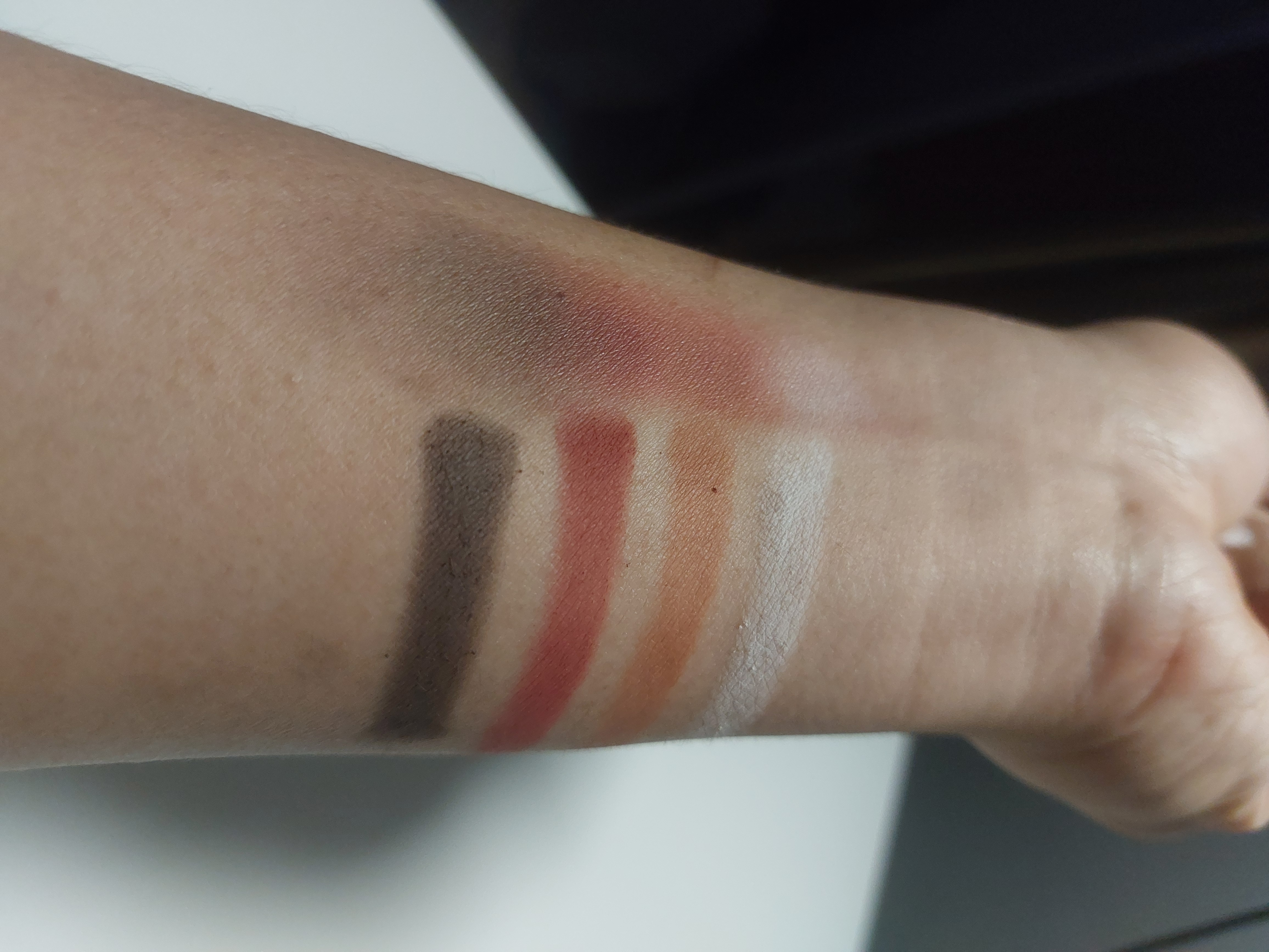 My arm with five eyeshadow swatches applied by brush on the forearm. The colors from top to bottom are: white, dark red, a muted red-brown, and a dark brown/black. The blending technique showcases the transitions between colors, revealing their blendability and pigmentation. The arm is positioned at an angle, and soft lighting highlights the texture and depth of the shades against the skin.