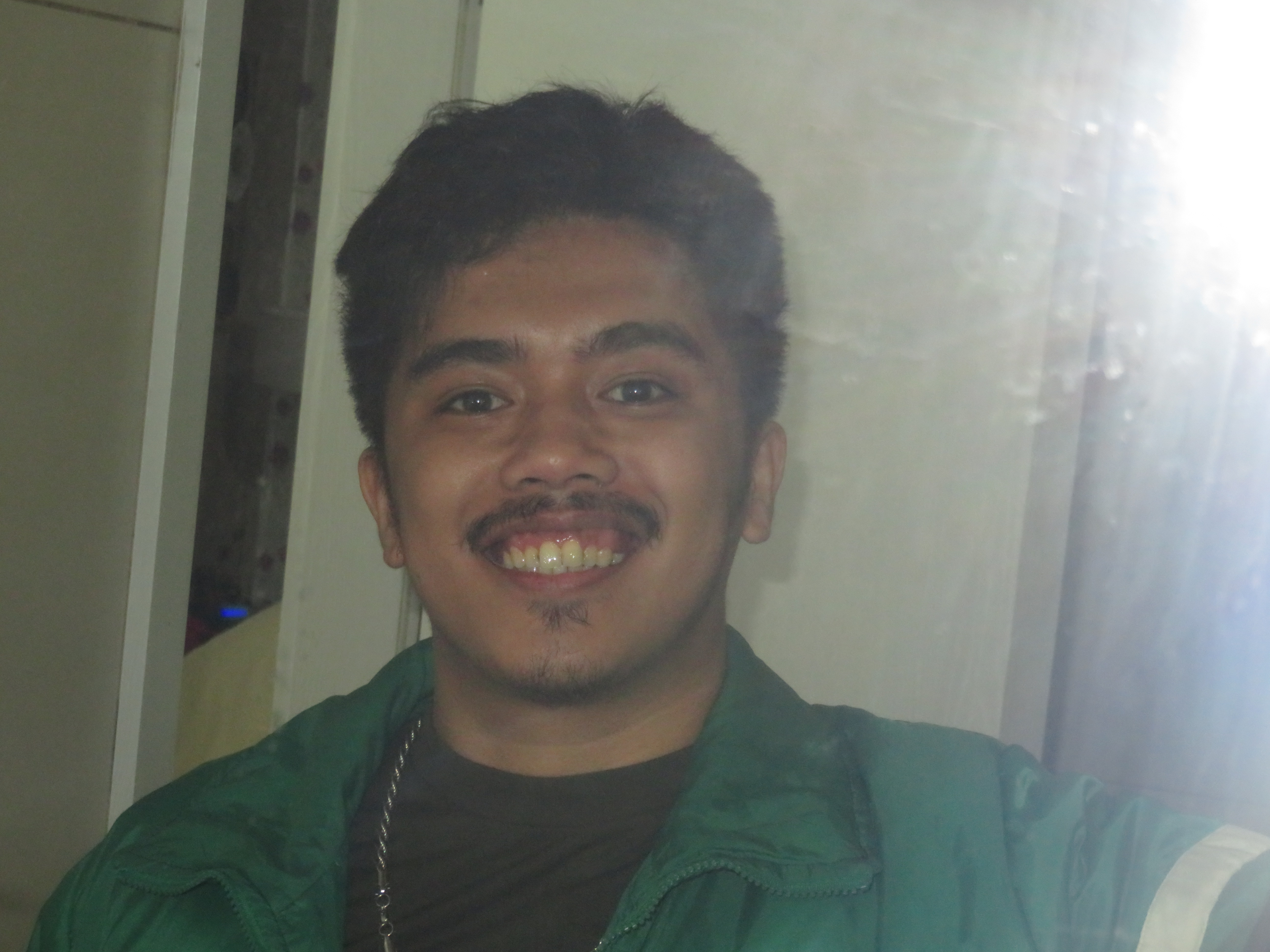 Me in a green jacket taking a mirror selfie using a digital camera with flash. I sport a wide smile showing my teeth.