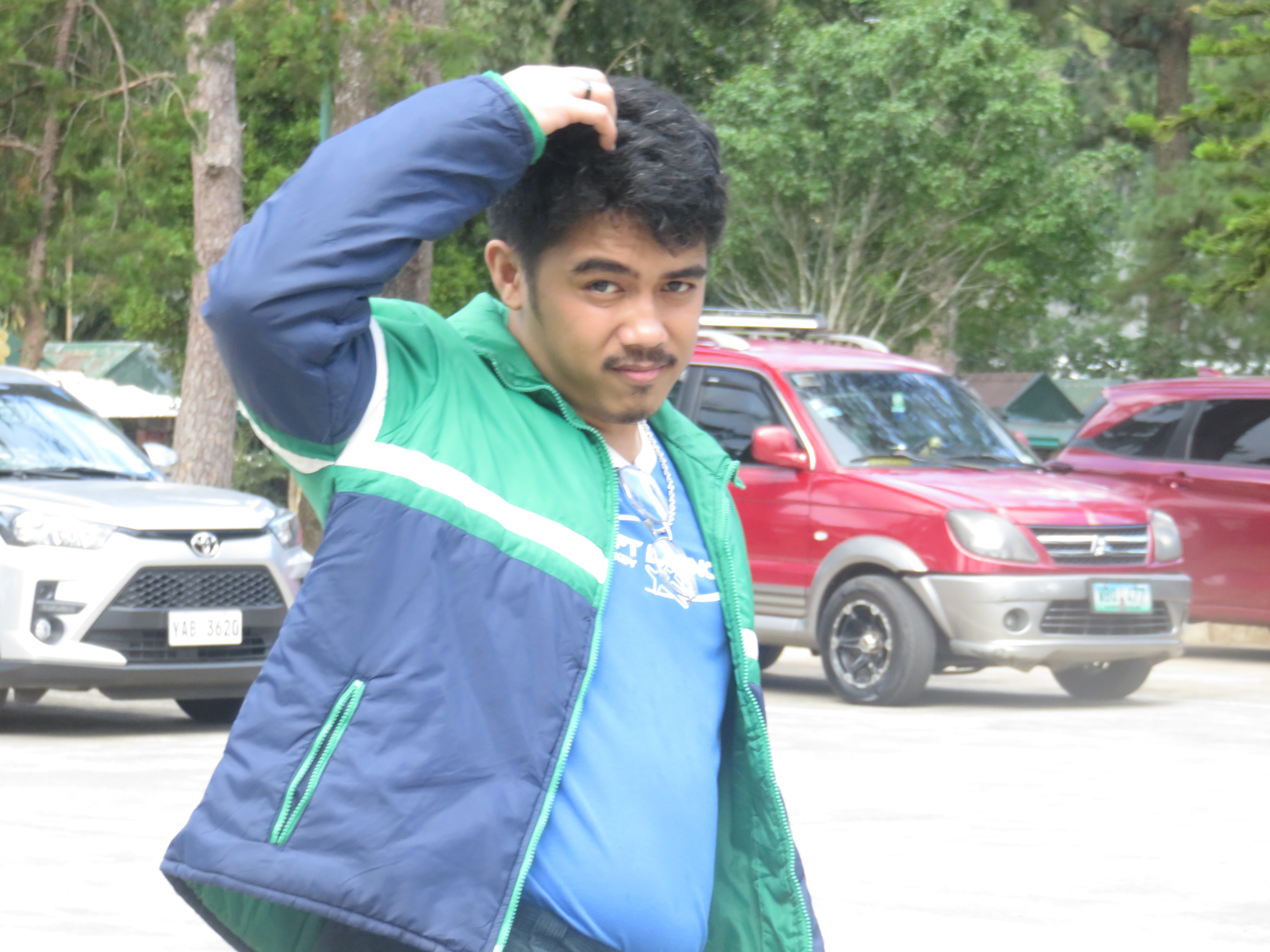 Me in a green jacket in a sunlit parking lot, scratching my head.