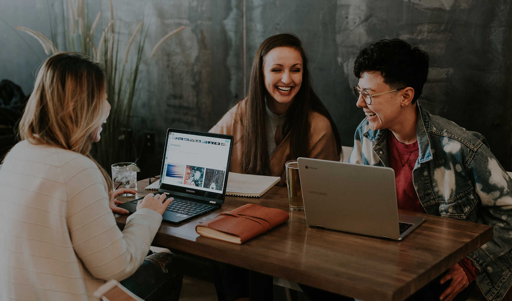 Three people sit at a table with laptops having a conversation.