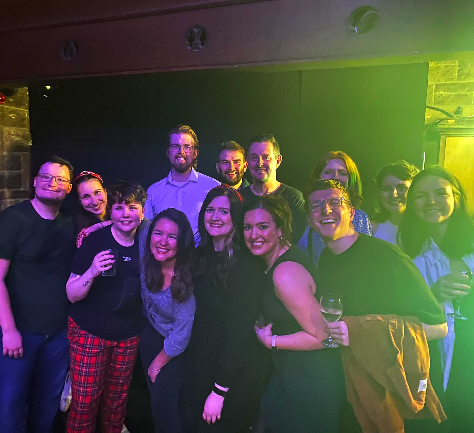Team 8MS pose for a group photo against a stone wall indoors, holding drinks illuminated with pink lights.