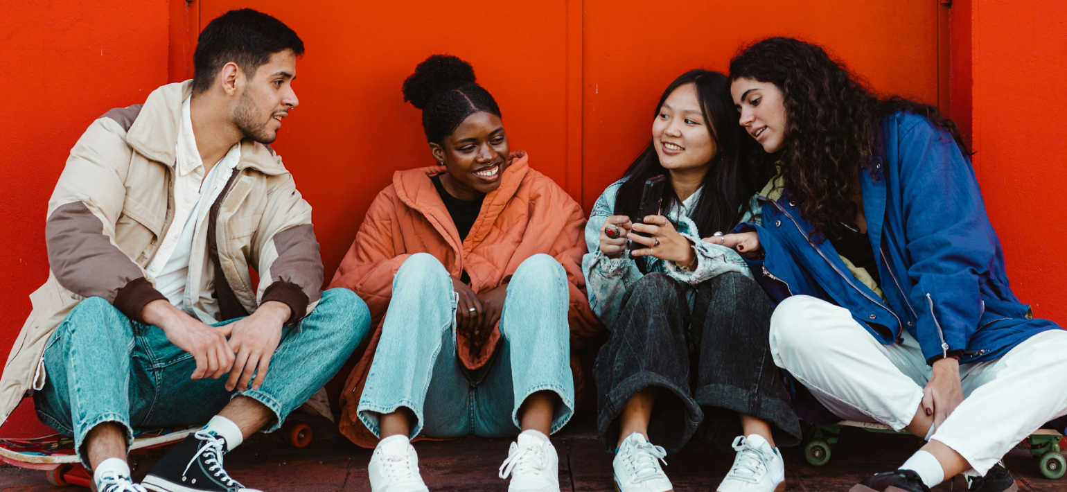 Four people sitting on a floor against a red background, looking at a phone.