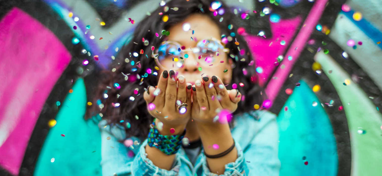 A dark-haired girl wearing glasses blowing confetti towards the camera against a colourful background.