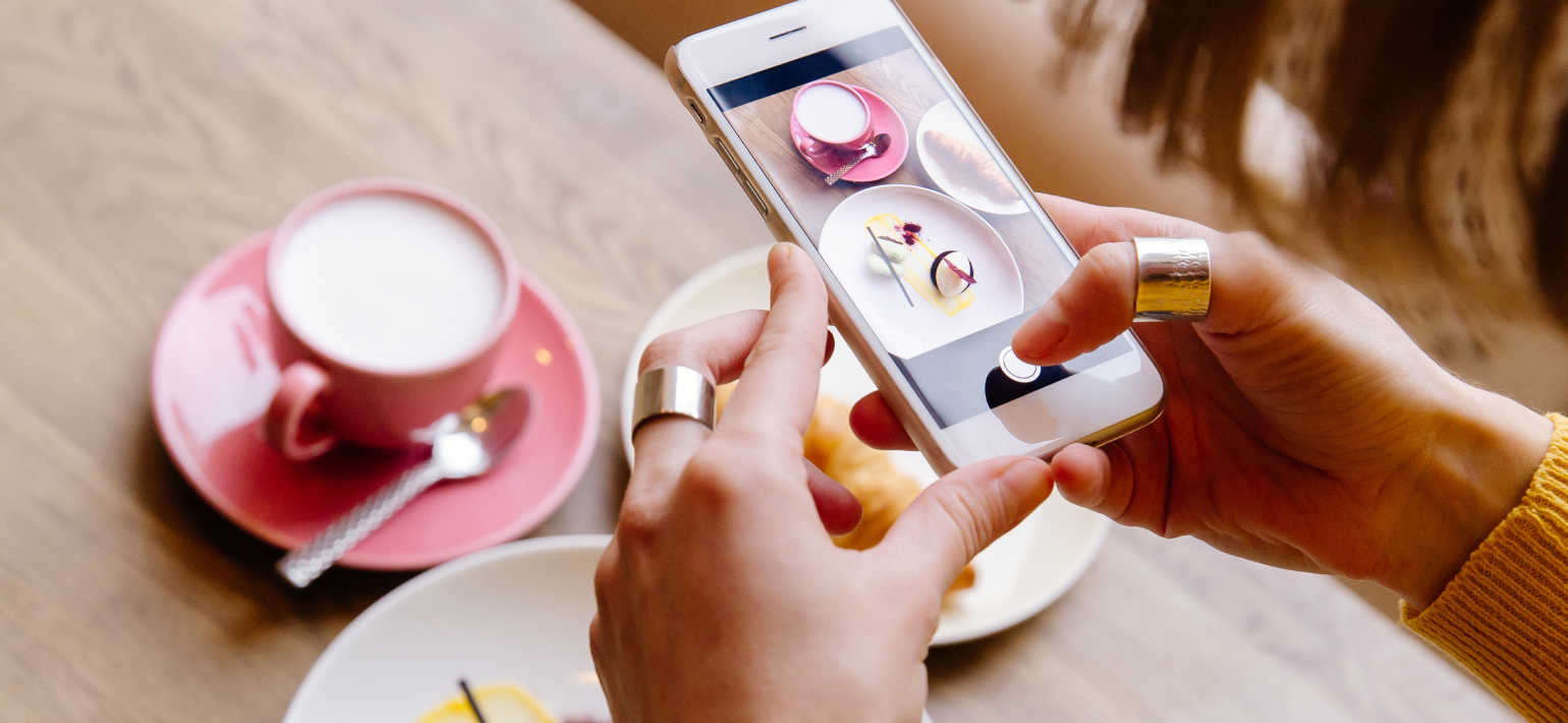 A person capturing an aesthetic photograph of coffee in a pink cup, next to a pastry and a cake.