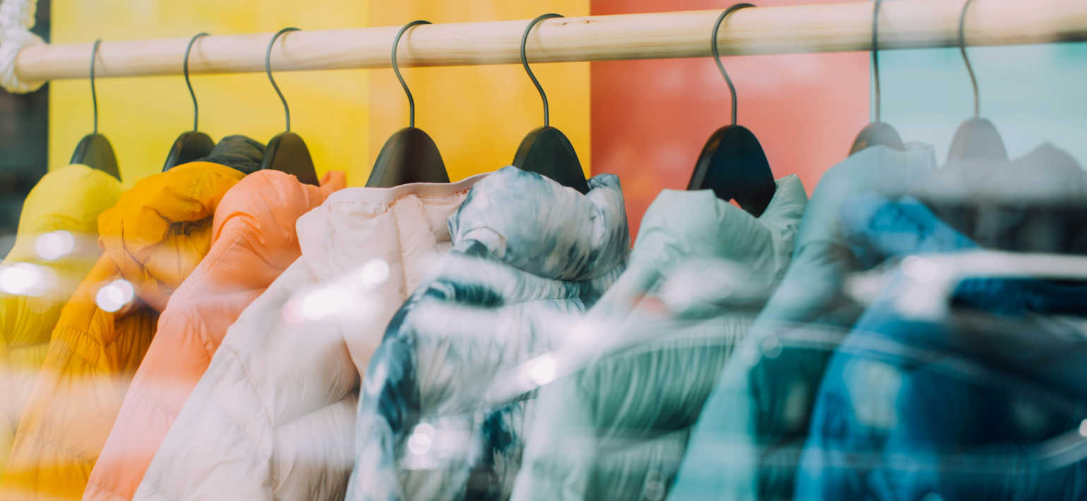 A row of puffer jackets hanging on black coat hangers from a wooden rail, set against a vibrant background.
