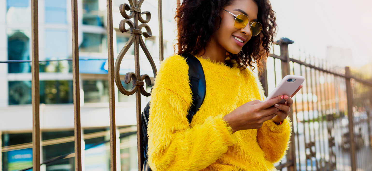 A woman stands in front of a bronze fence wearing a fluffy yellow jumper and yellow-tinted glasses, looking at her phone.