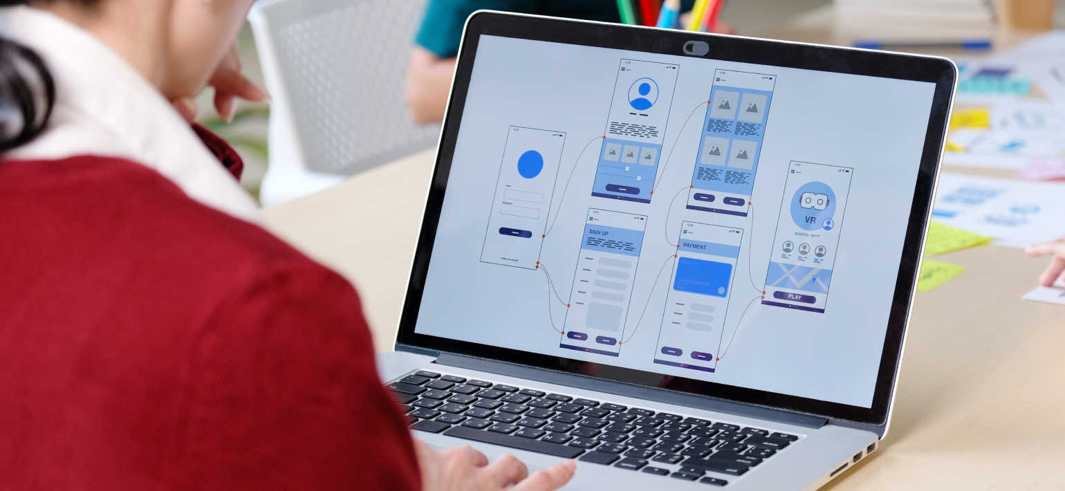 A person wearing a red jumper looks at a laptop with a diagram of an app infrastructure on the screen.