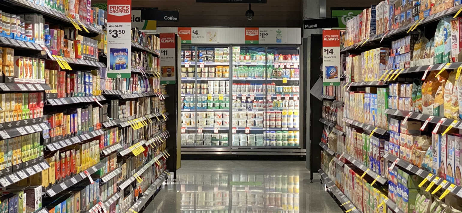 A fully stocked isle in a supermarket with a fridge at the end displaying dairy products.