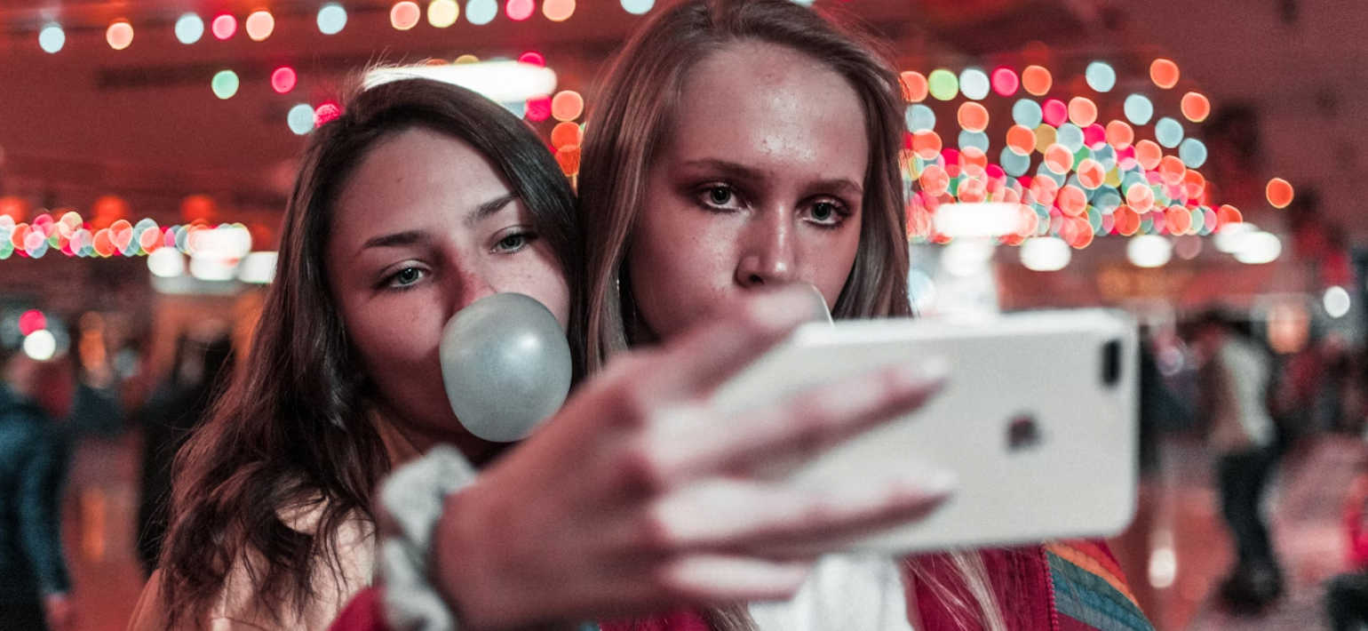 Two woman take a selfie while one blows a bubble with bubblegum.