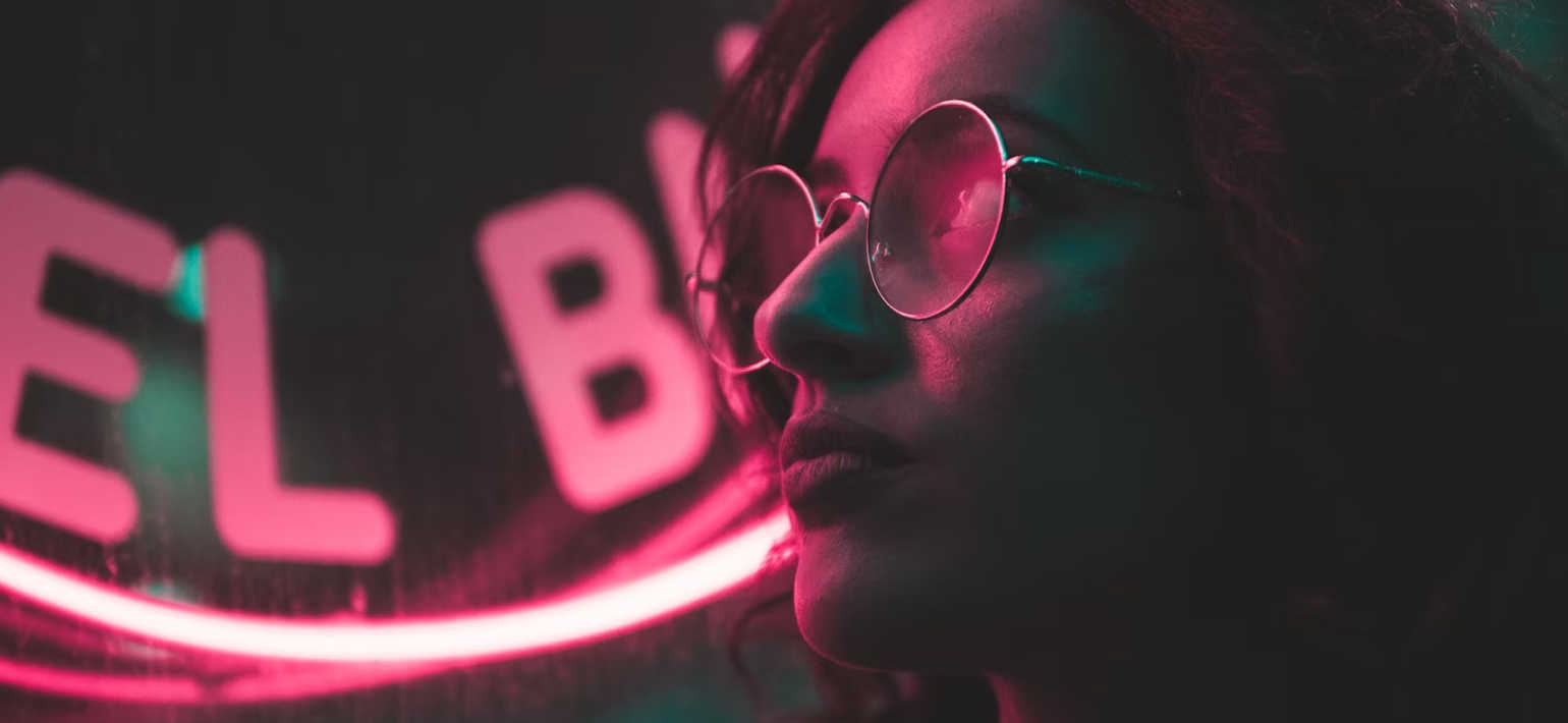 A person wearing round glasses in front of a pink neon sign.