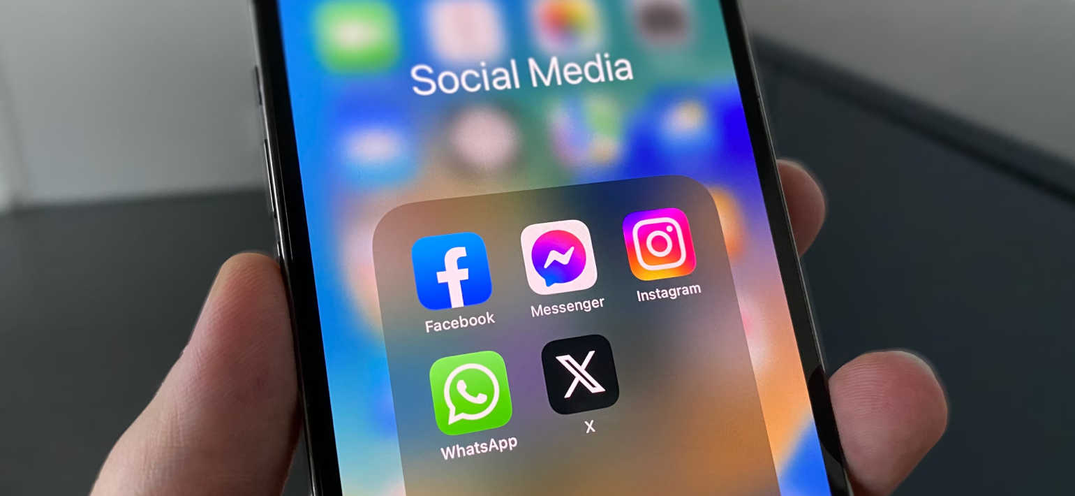 A close up of a phone on the with a 'social media' folder showing app icons for FaceBook, Messenger, Instagram, WhatsApp and X.