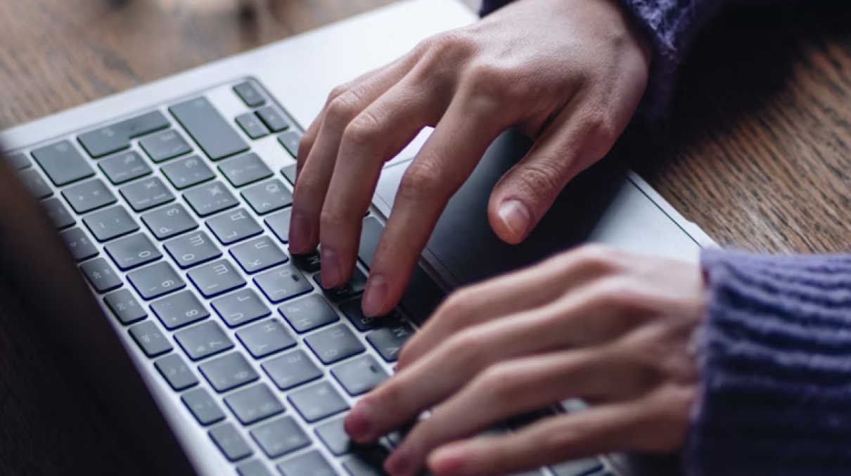 The hands of a person wearing a sweater with purple cuffs, typing on an Apple Macbook