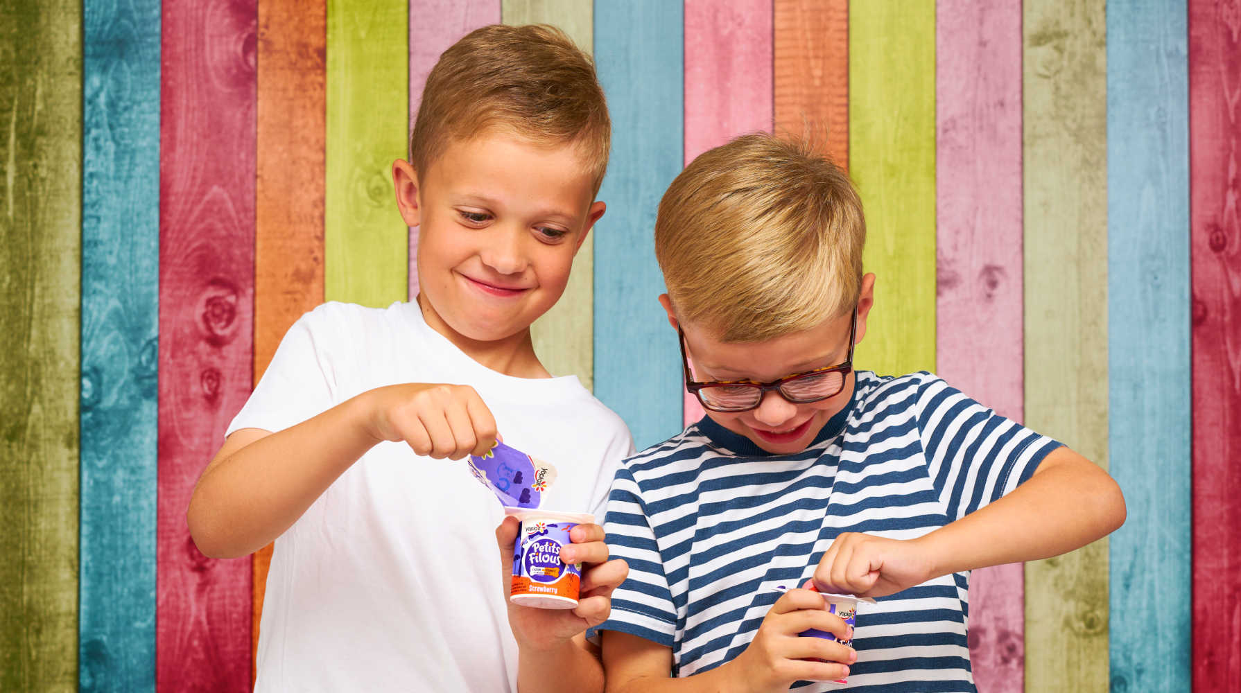 Two young boys open Petits Filous yoghurts in front of a colourful wooden panel background.