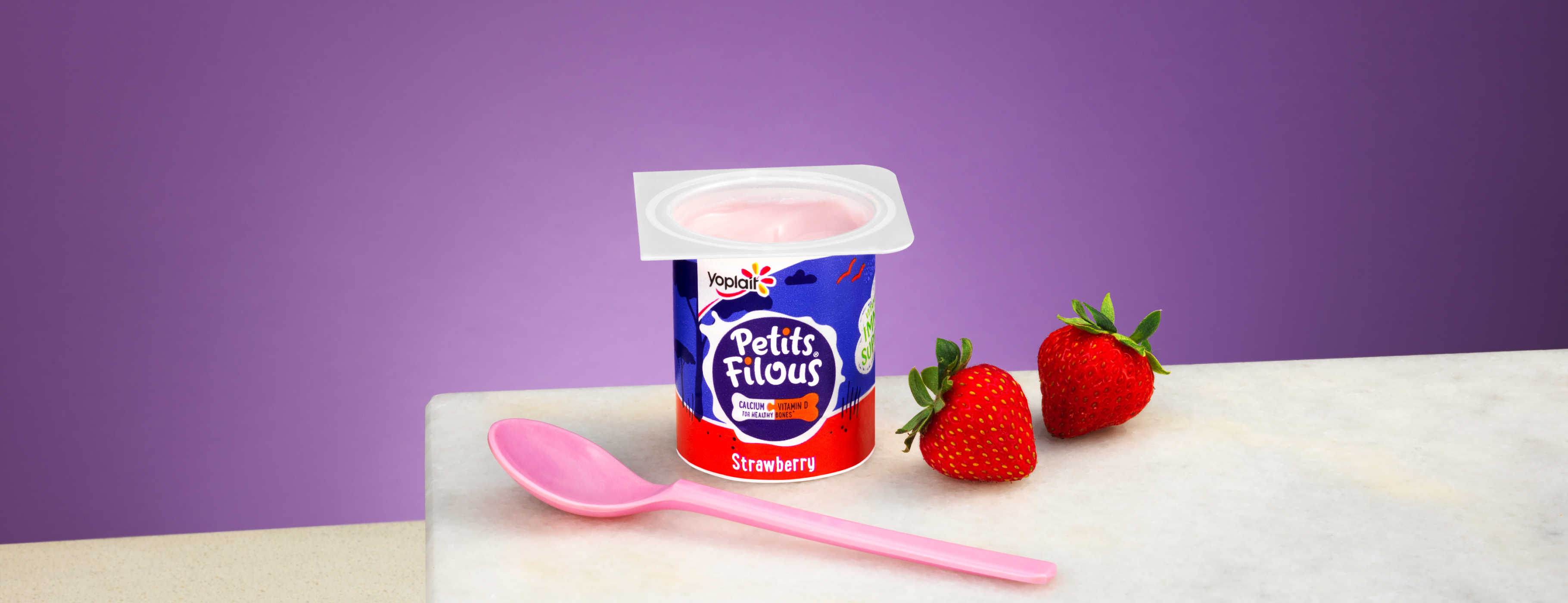 A strawberry Petits Filous yoghurt pot is shown with a pink spoon and two strawberries on a marble surface against a purple background.