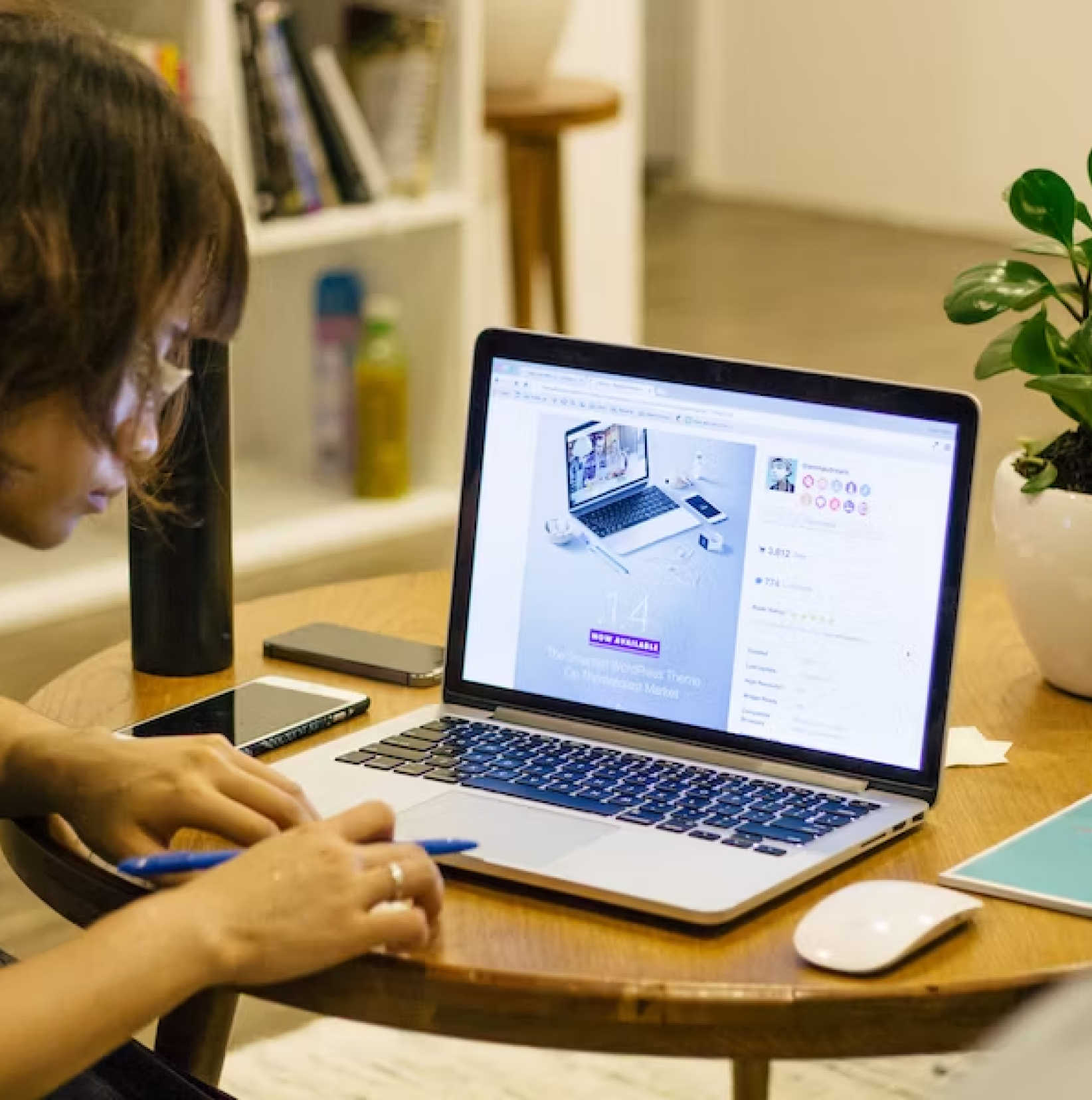 A person works at a table with a laptop, jotting notes beside a smartphone and potted plant.