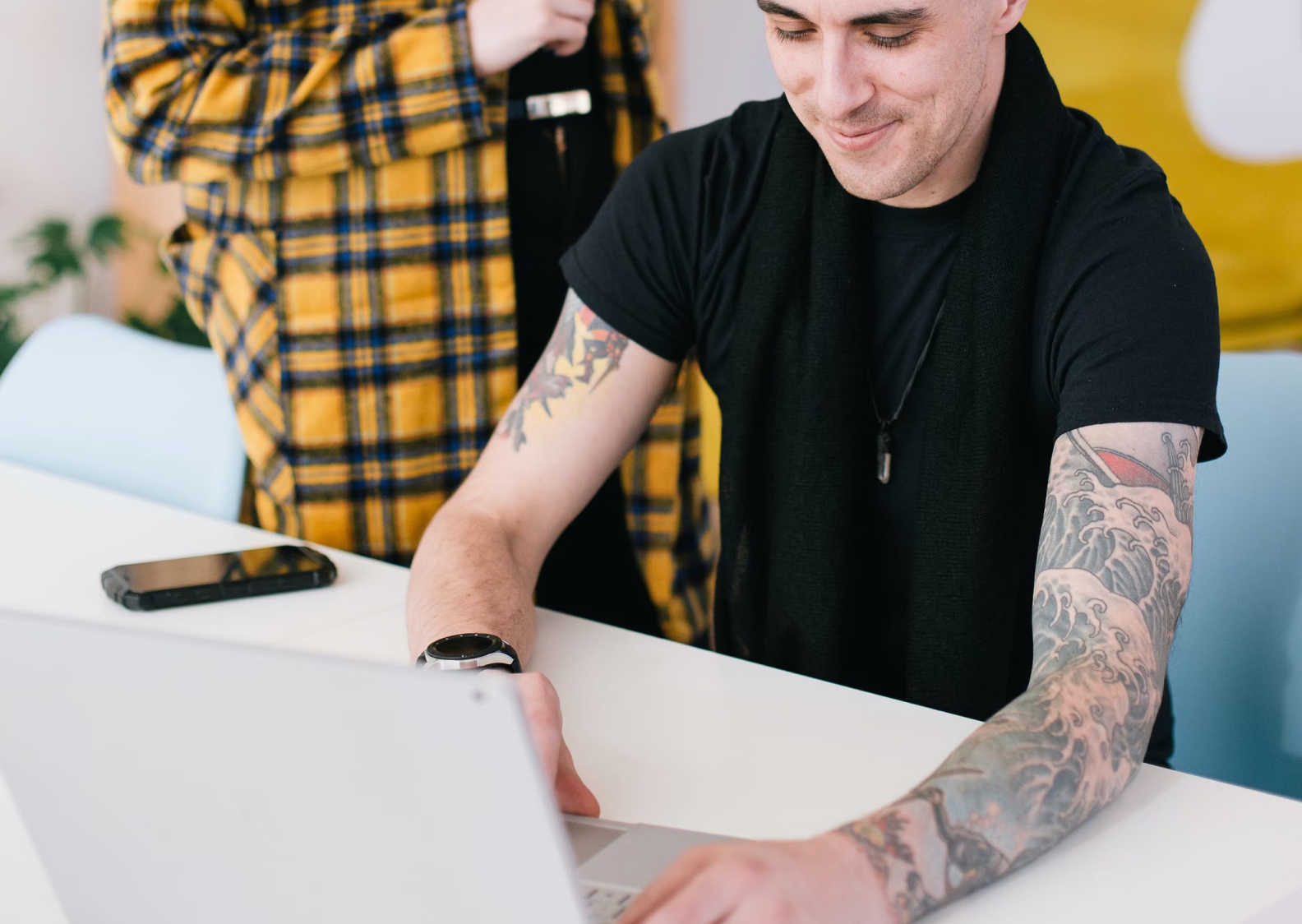 A man with tattooed arms is smiling while working on a laptop at a white desk.