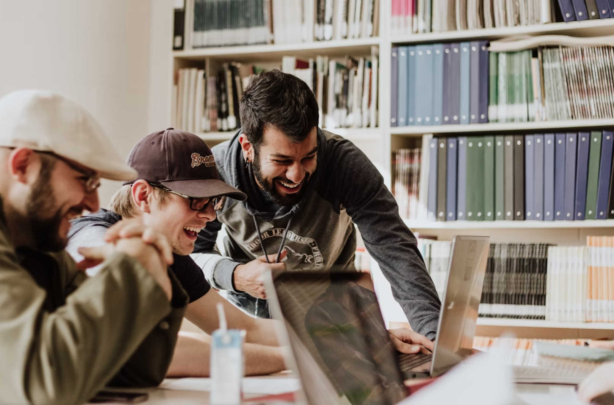 A group of people laugh and collaborate around laptops in a study room.