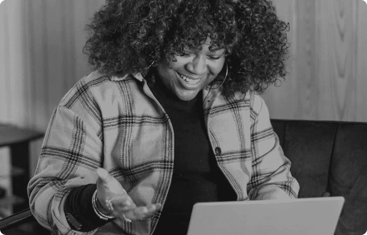 A woman sits on a sofa smiling and gesturing while looking at a laptop.