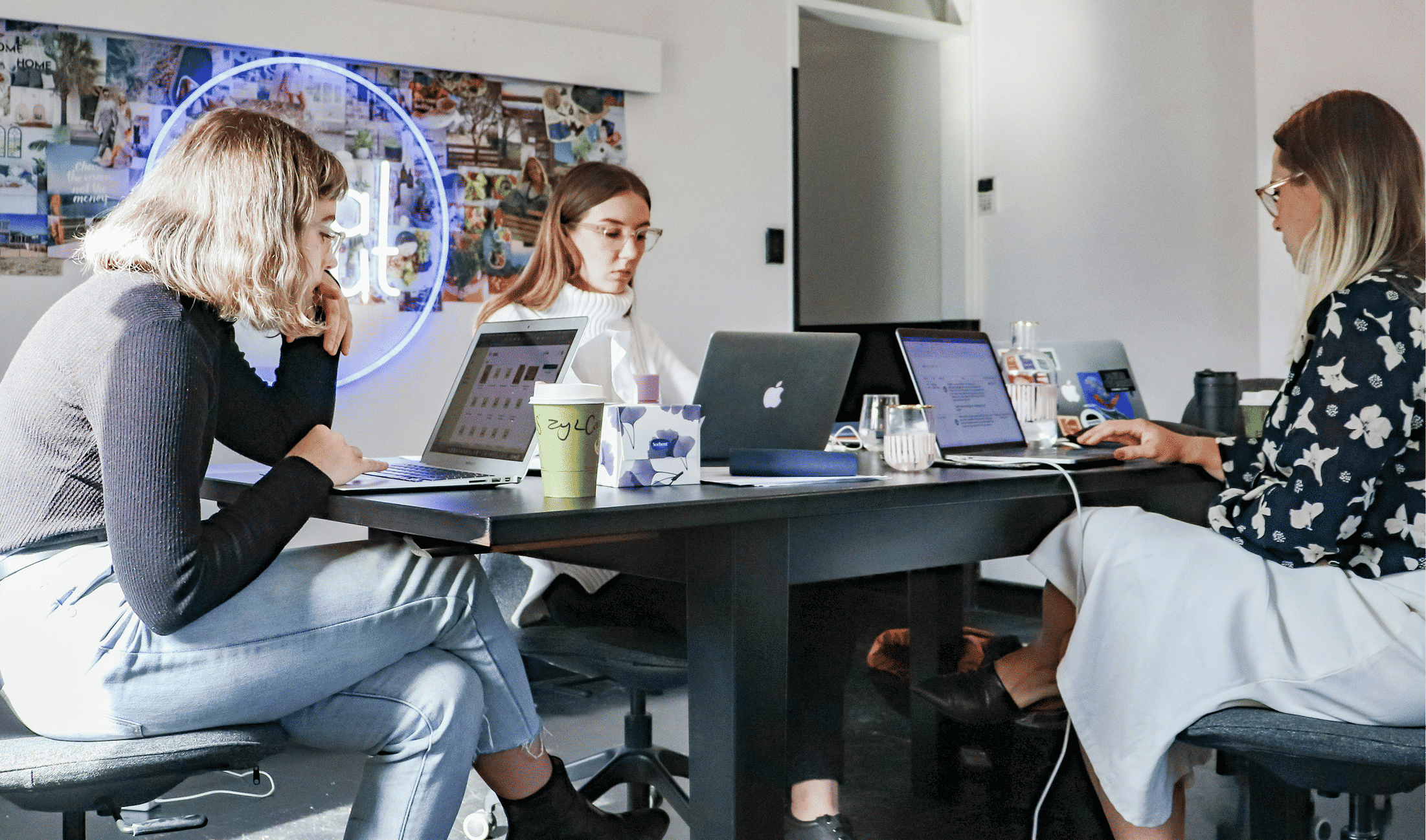 Three women working on laptops around a table in an office setting.