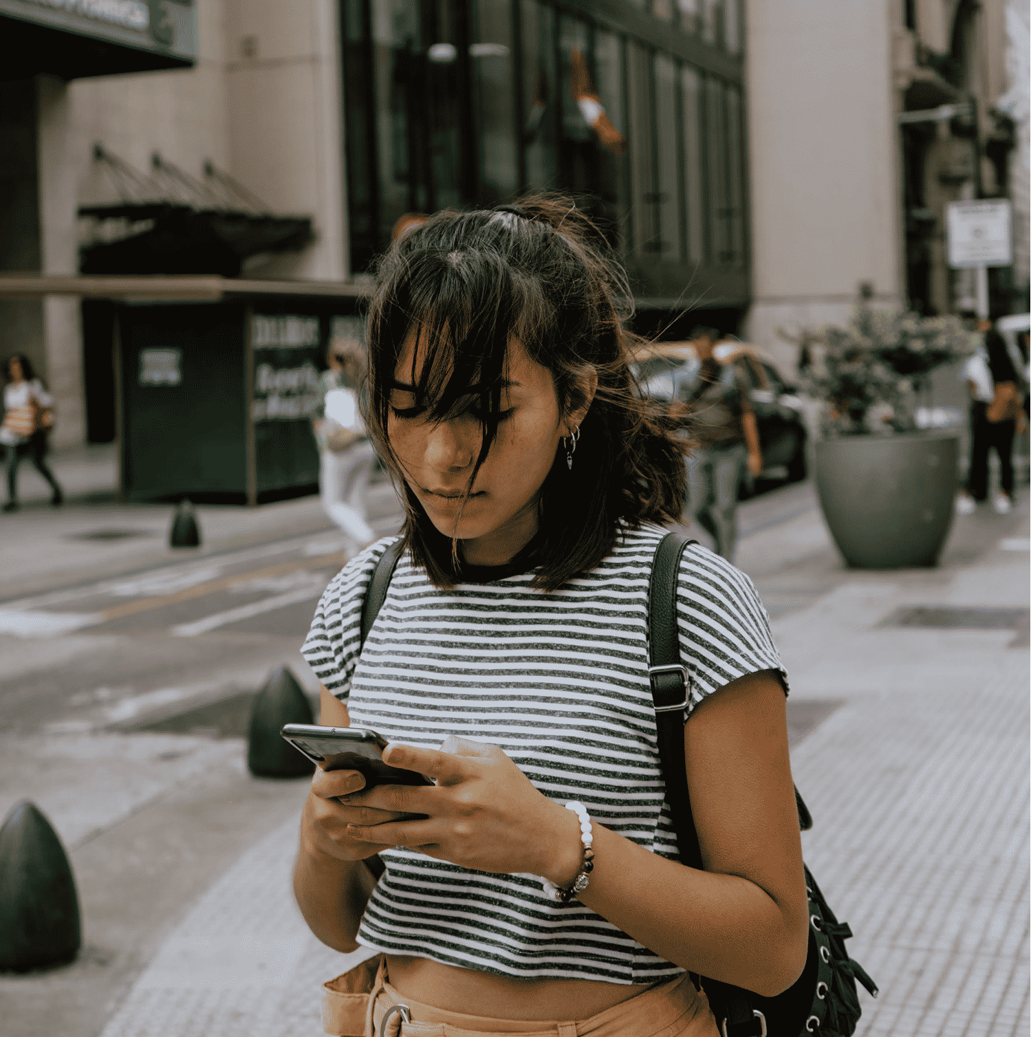 A woman wearing a striped top and yellow trousers is walking on a city street while looking at her smartphone.