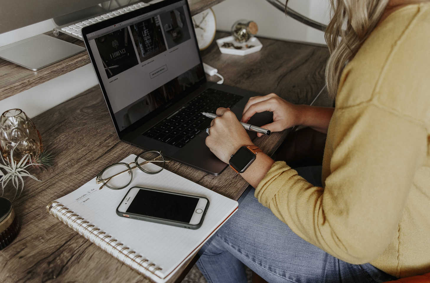 A woman working at a desk with a laptop, smartphone, notebook, and glasses.