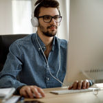 A young male with glasses wearing headphones using the mouse looking at his desktop screen