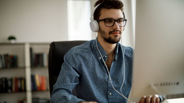 A young male with glasses wearing headphones using the mouse looking at his desktop screen