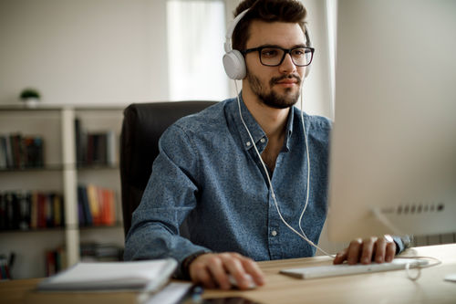 A young male with glasses wearing headphones using the mouse looking at his desktop screen