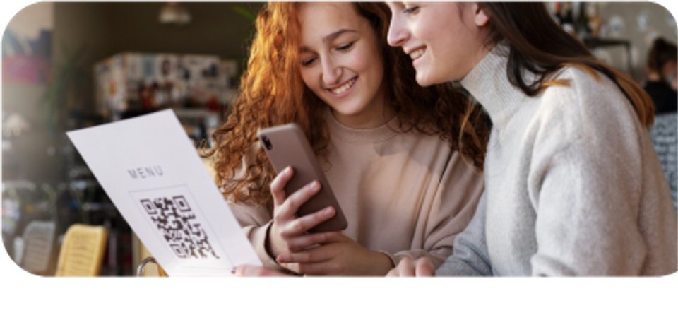 Two girls are sitting at the restaurant, one of them looks at the menu, the other one on her smartphone.