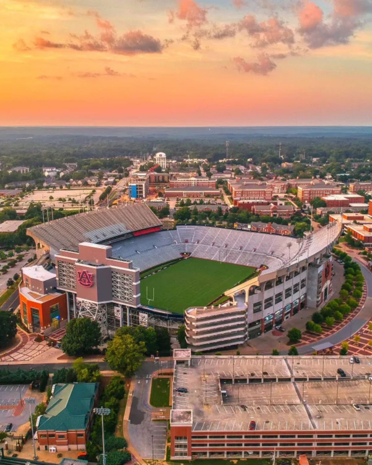 Aerial view of Auburn University’s football stadium in Auburn, Alabama, showing the oval stands surrounding the green field with campus buildings and tree-lined streets stretching into the distance at sunset.
