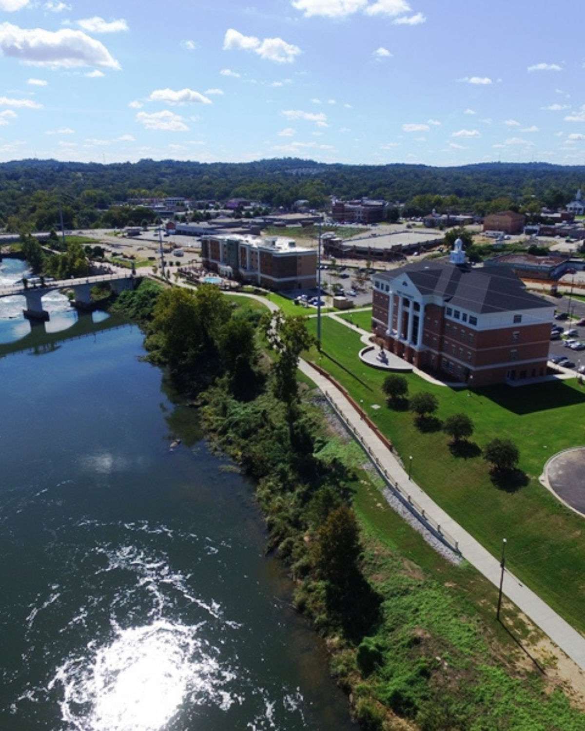 Aerial view of the Phenix City Riverwalk in Phenix City, Alabama, with a paved riverside trail and green embankment running alongside the water, passing a large brick civic building and nearby downtown streets and bridges.
