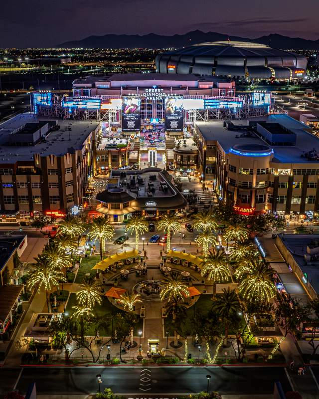 Nighttime view of Glendale, Arizona’s entertainment district, with palm trees wrapped in lights leading to a brightly lit plaza of shops and restaurants, and the Desert Diamond Arena in the background.