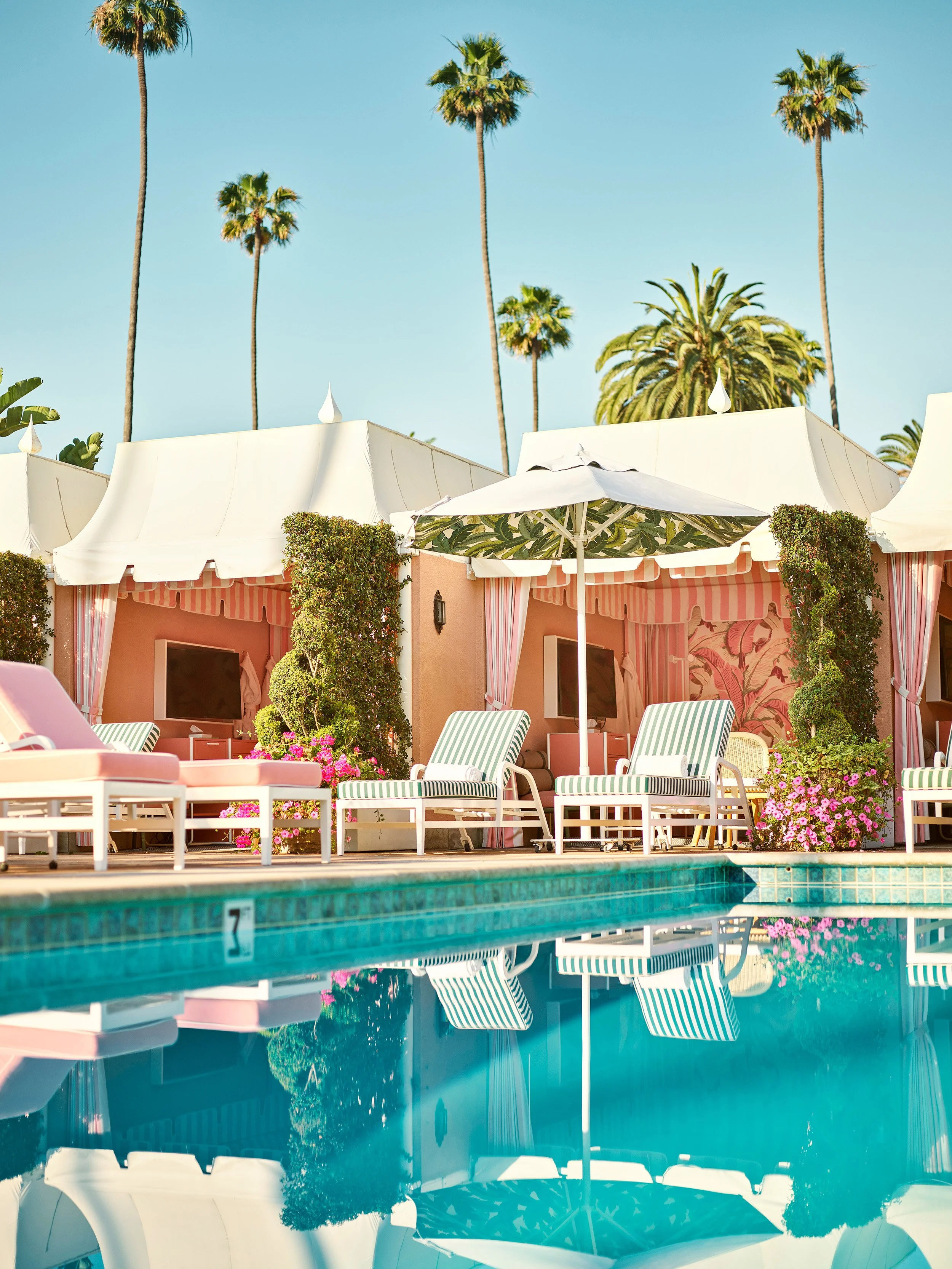 Iconic pool at The Beverly Hills Hotel in Beverly Hills, California, surrounded by striped lounge chairs, pink cabanas, palm trees, and tropical landscaping.