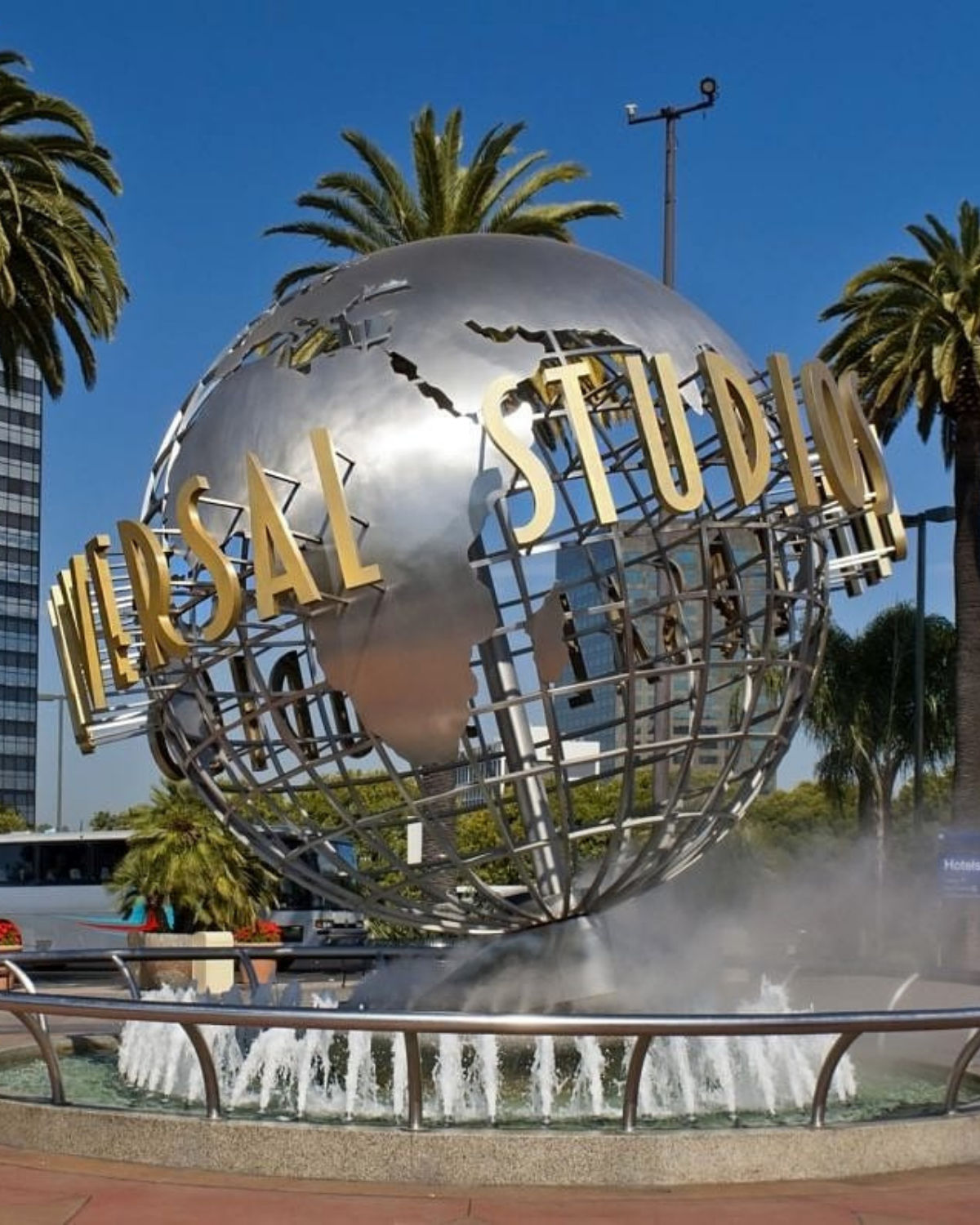 Close-up of the Universal Studios globe fountain near Burbank, California, with the metallic rotating globe, bold gold “Universal Studios” lettering, and palm trees in the background.