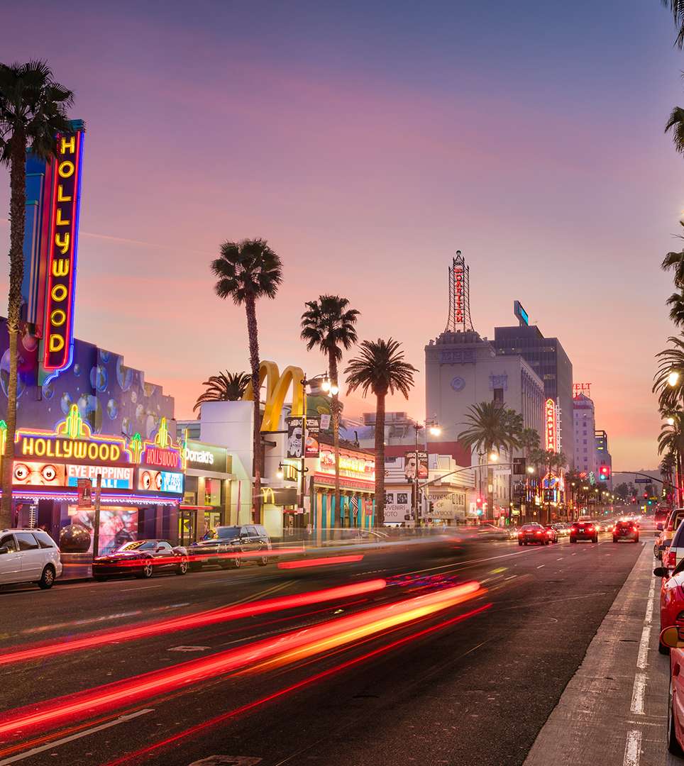 Hollywood Boulevard at sunset with colorful neon signs, palm trees, storefronts, moving traffic, and illuminated buildings in the greater Los Angeles area near Glendale, California.