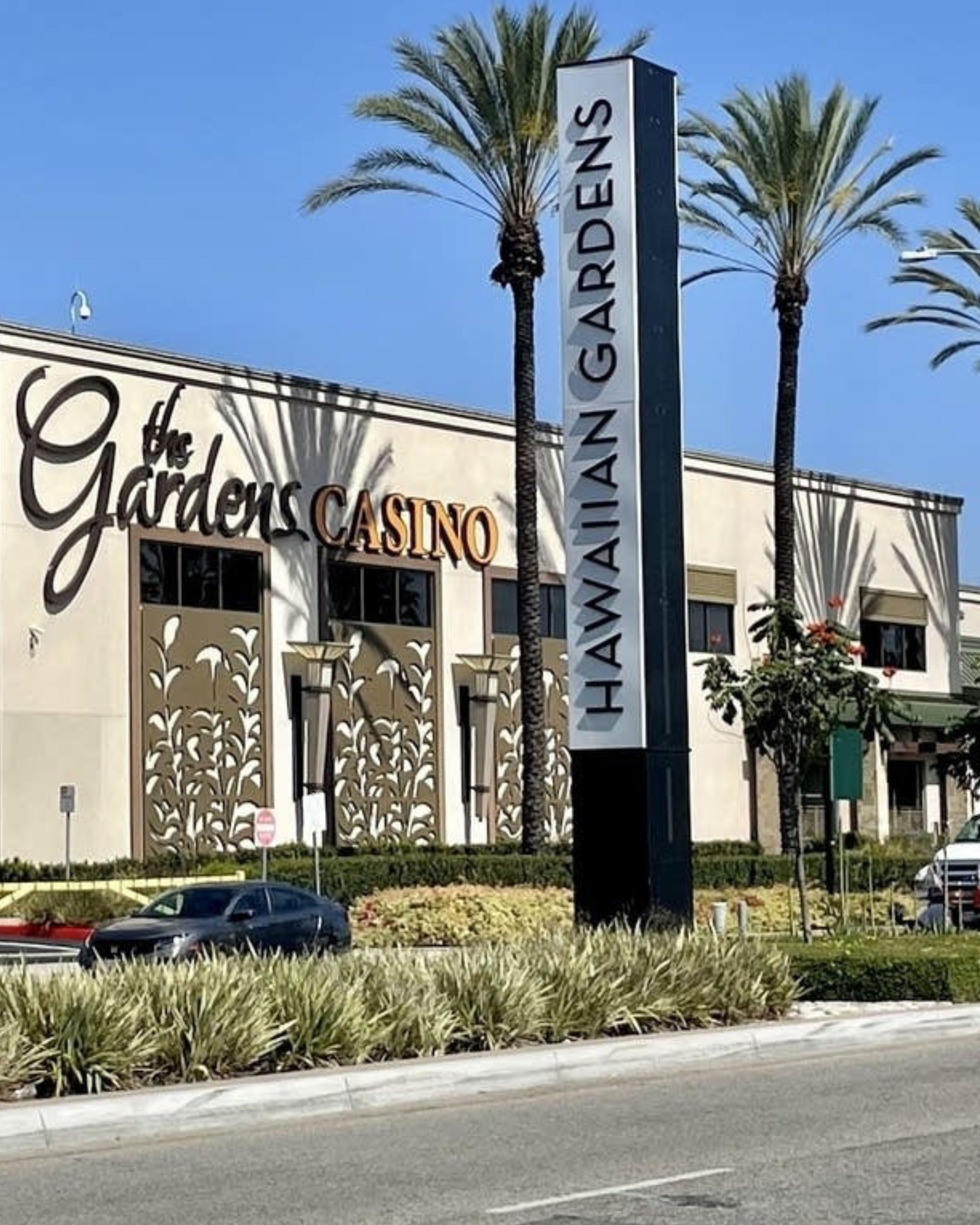 Exterior of the Gardens Casino in Hawaiian Gardens, California, featuring the large Hawaiian Gardens sign, casino building, tall palm trees, landscaping, and street in the foreground.