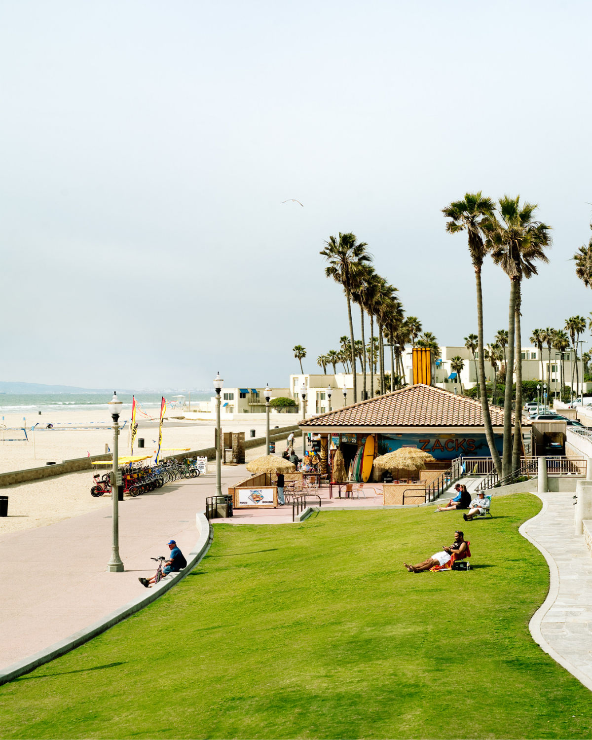 Daytime scene at Huntington Beach, California, showing a grassy bluff with people relaxing, a beachfront path lined with lampposts, tall palm trees, and a small café building beside the sandy beach and ocean.