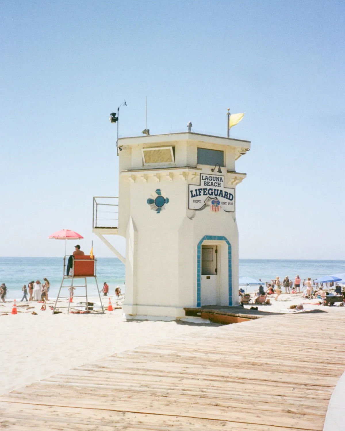 Sunny beach scene in Laguna Beach, California, featuring the iconic white lifeguard tower on the sand beside a wooden boardwalk, with a lifeguard stand, beachgoers, and the blue ocean in the background.