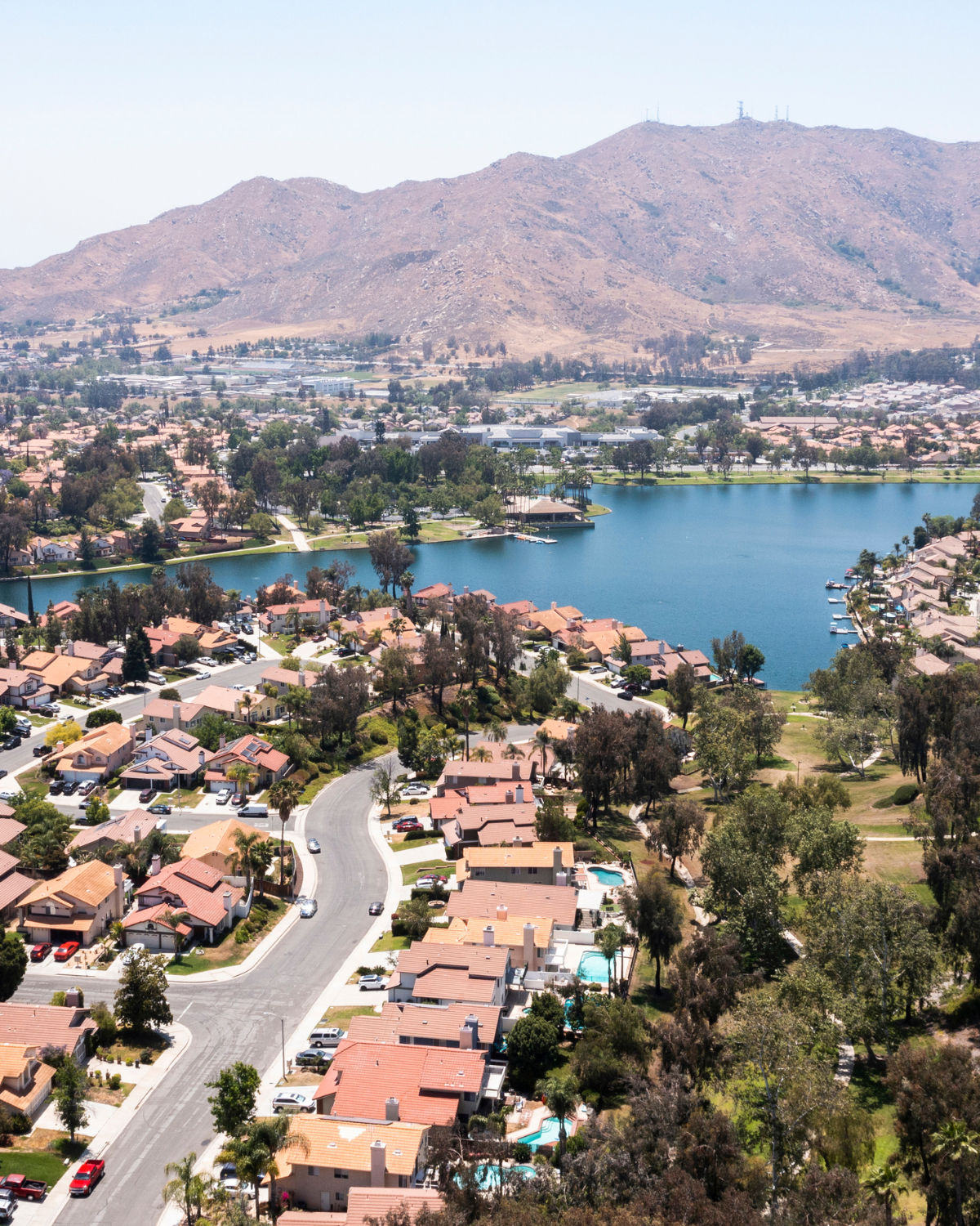 Aerial view of a calm blue lake surrounded by red-tile-roofed homes, green trees, and golden hills in Moreno Valley, CA