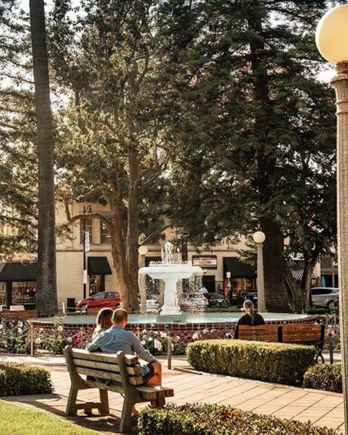 Afternoon scene in Old Towne Orange, California, showing a couple sitting on a bench facing a white fountain in a tree-shaded plaza, with neatly trimmed hedges, flowerbeds, and historic storefronts in the background.