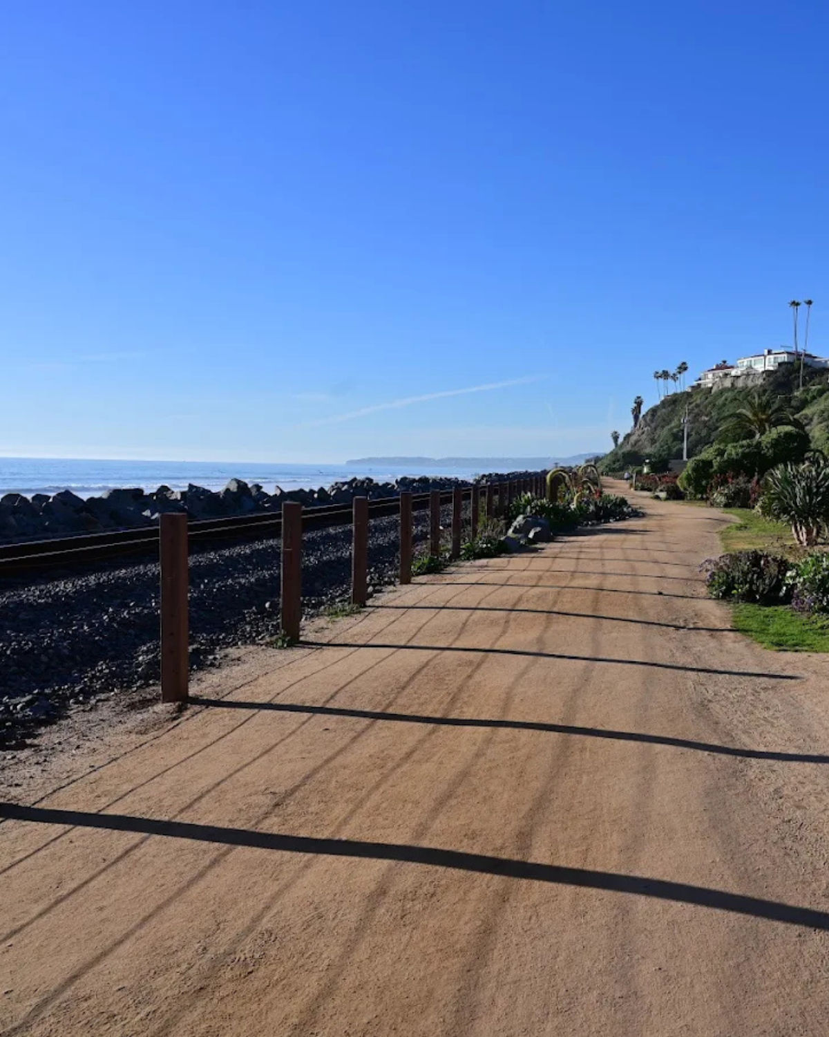 Scenic view of the San Clemente Beach Trail in San Clemente, California, with a wooden boardwalk curving along the shoreline beside green bluffs, the ocean to the left, and birds flying under a dramatic blue and cloud-filled sky.