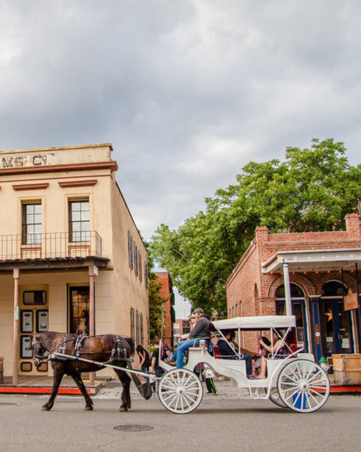 Street scene at the Old Sacramento Waterfront in Sacramento, California, showing a horse-drawn white carriage passing historic brick and stucco buildings, with people riding inside under a cloudy sky.