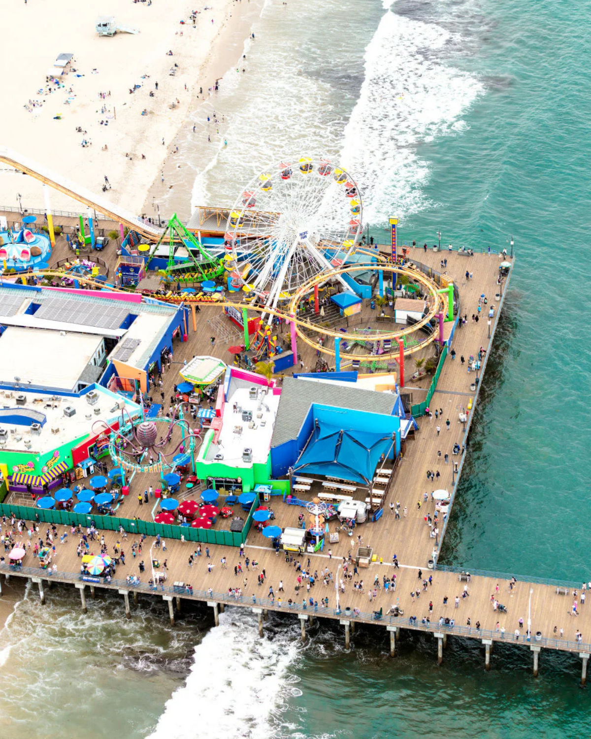 Aerial view of the Santa Monica Pier in Santa Monica, California, showing the colorful amusement park with its Ferris wheel and roller coaster, crowds on the wooden pier, and waves from the Pacific Ocean crashing below and onto the adjacent sandy beach.