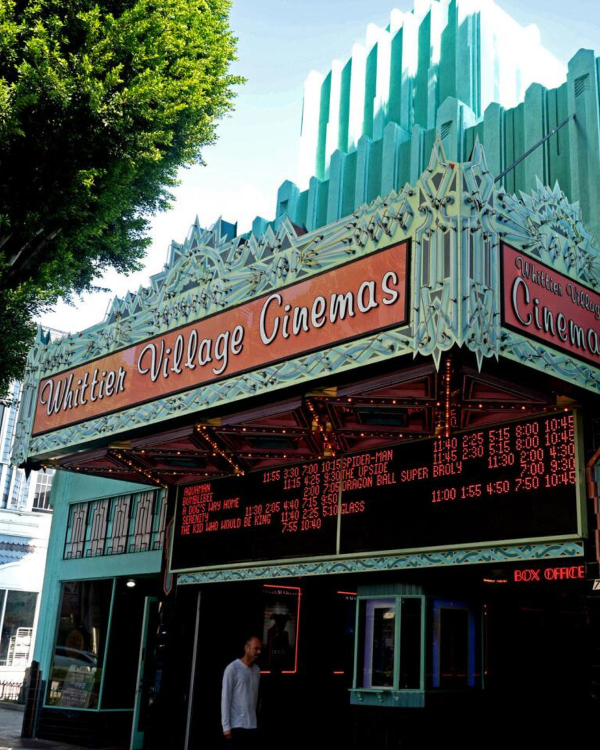 Street-level view of Whittier Village Cinemas in Whittier, California, showcasing the theater’s ornate Art Deco marquee with its bright movie listings and turquoise facade beneath leafy trees.