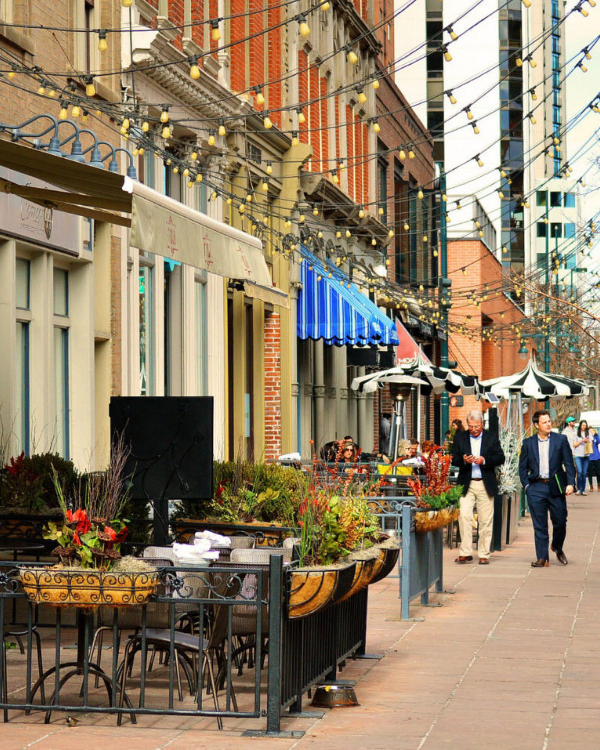 Downtown street scene in Arvada, Colorado, featuring a row of brick and historic-style buildings with sidewalk patios full of planters, string lights crisscrossing overhead, and people walking along the wide pedestrian-friendly walkway.