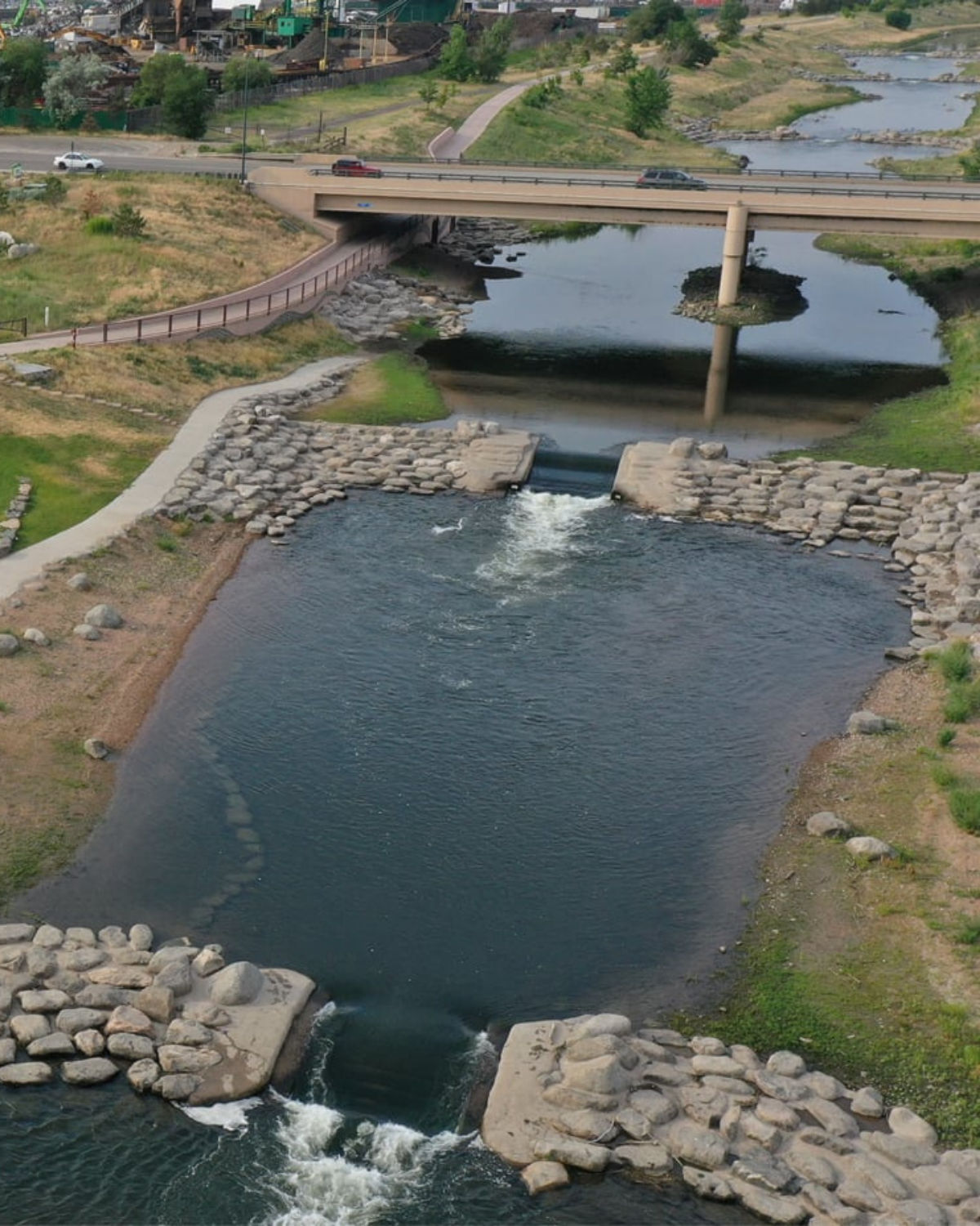 Aerial view of River Run Park in Englewood, Colorado, showing a section of the South Platte River flowing through rock-lined chutes, with walking paths on both banks and a bridge carrying a road over the water.